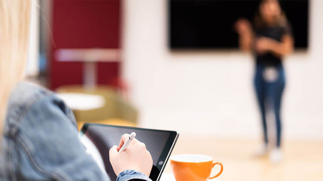 Une femme blonde portant un blouson en jean bleu prend des notes sur une tablette noire. Elle est assise devant une tasse à café orange. Elle regarde une présentation sur un grand écran.