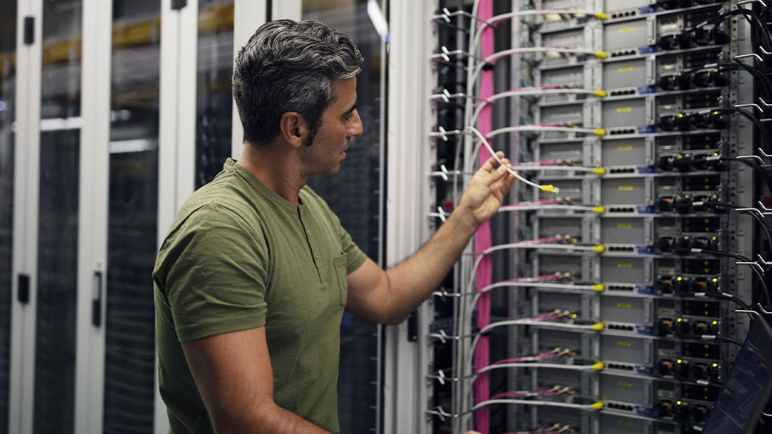 Mann mit kurzen schwarzen Haaren trägt ein grünes T-Shirt und steht vor einem grossen Computer. Dort hantiert er mit einem Internet-Kabel.