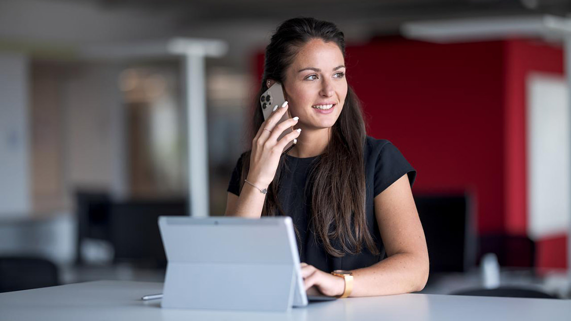 La photo montre une jeune femme qui téléphone via son smartphone, avec devant elle une tablette sur le bureau. 