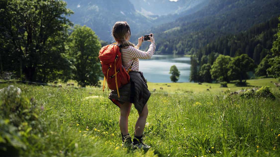 Una donna con uno zaino da montagna rosso in un paesaggio alpino è in piedi davanti a un lago. Ha uno smartphone in mano e lo usa per scattare una foto