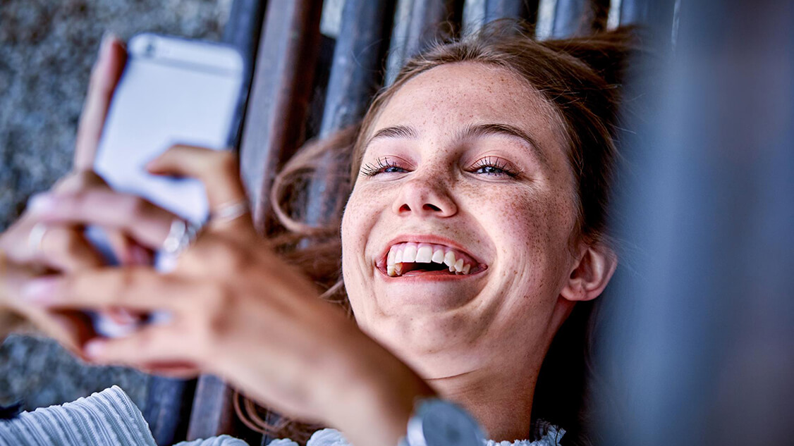 Auf dem Bild ist eine Frau zu sehen, die lachend einen Inhalt auf einem Smartphone betrachtet.