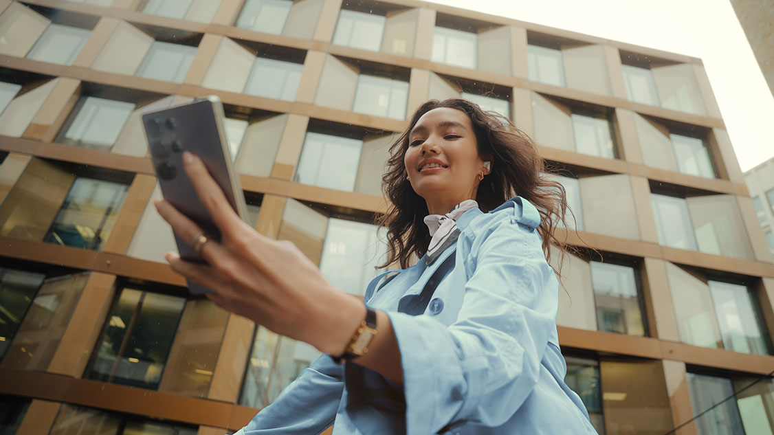 A young woman in a light blue coat holds a smartphone in her hand and smiles while standing outside in front of a modern building.