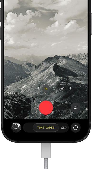 iPhone 18 Pro mostrato di fronte, sul display un paesaggio montuoso, con fotocamera in modalità Time-Lapse e cavo di alimentazione collegato alla porta di ricarica
