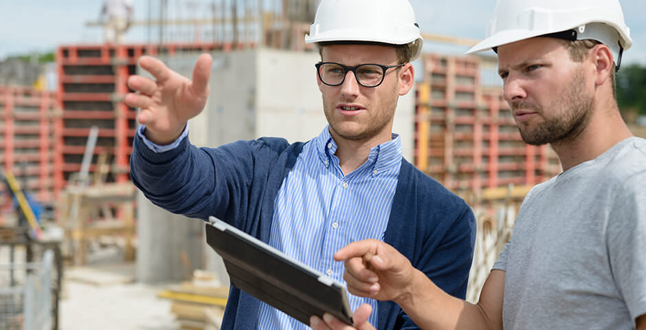 Zwei Männer mit Sicherheitshelmen auf Baustelle. Sie sprechen zusammen, zeigen mit den Händen in die Ferne.