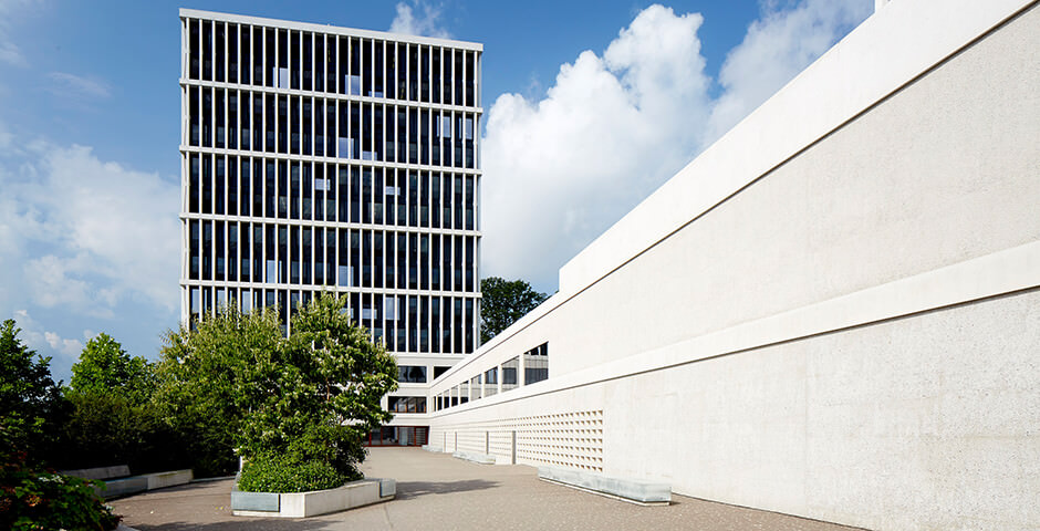 L’image montre le bâtiment du Tribunal administratif fédéral à Saint-Gall. Le ciel est bleu avec des nuages blancs et le bâtiment est gris/blanc. Des arbres et des buissons sont visibles devant le bâtiment. Le bâtiment lui-même est un immeuble moderne avec beaucoup de grandes fenêtres.