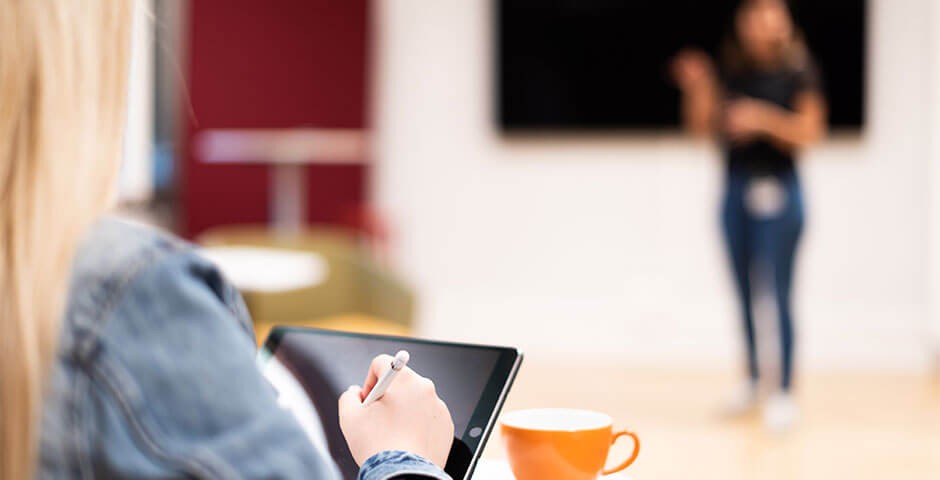 Une femme blonde portant un blouson en jean bleu prend des notes sur une tablette noire. Elle est assise devant une tasse à café orange. Elle regarde une présentation sur un grand écran.