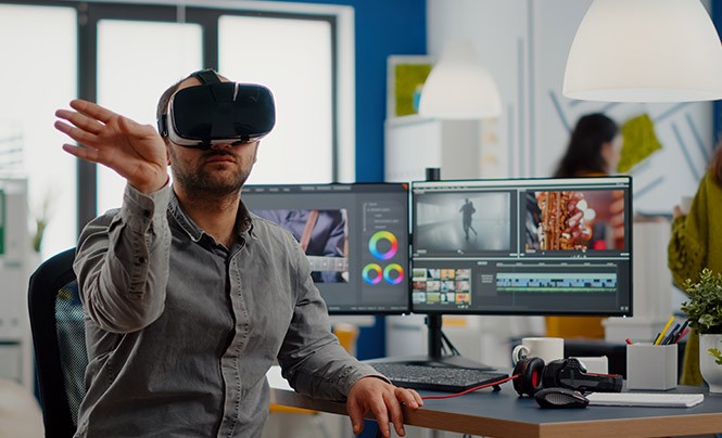 Man in a grey shirt sits in an office in front of 2 monitors that are running a video-cutting program. He is wearing a virtual reality headset and is pointing to the right with his right hand.